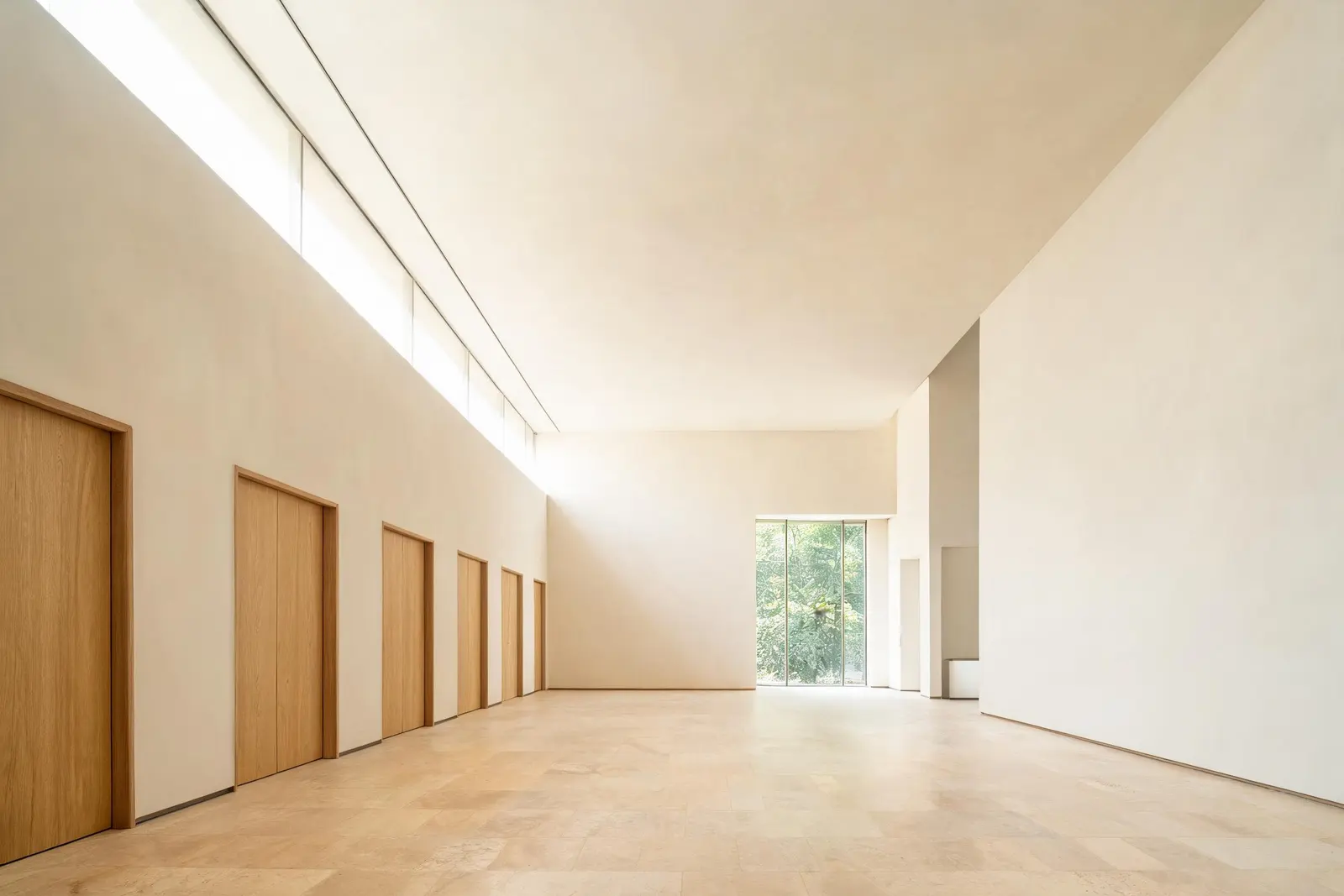 La galleria interna del poliambulatorio: alte porte in rovere a filo muro, pavimento in travertino chiaro e una fessura continua di luce naturale lungo il soffitto.