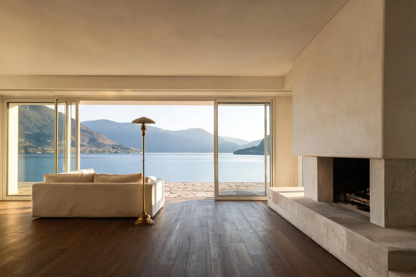 Soggiorno di una villa sul Lago Maggiore: vetrate scorrevoli a tutta altezza aperte sulla terrazza e sul lago, pareti in intonaco chiaro, pavimento in rovere scuro e camino in pietra.