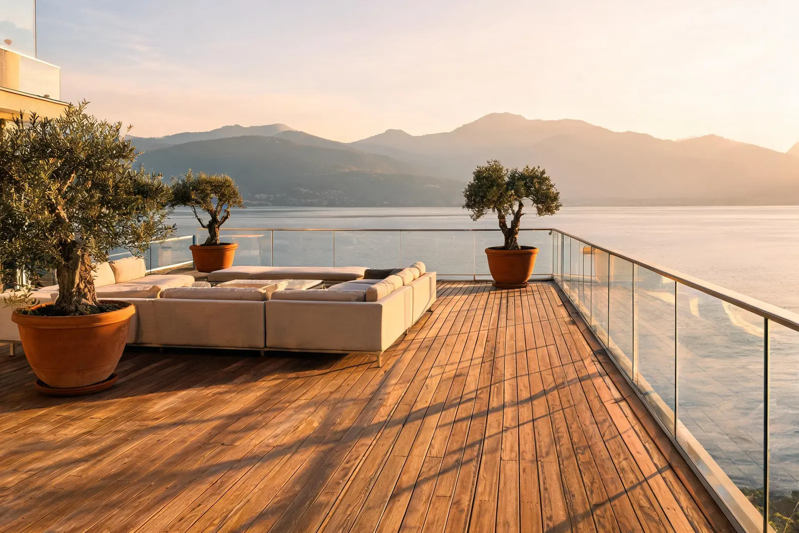 Attico Riva a Locarno: terrazza sul tetto con pavimento in teak, sedute basse in lino, parapetto in acciaio e vetro e ulivi in vaso davanti al lago.